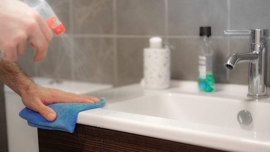 Person uses spray and blue microfibre cloth to clean bathroom sink
