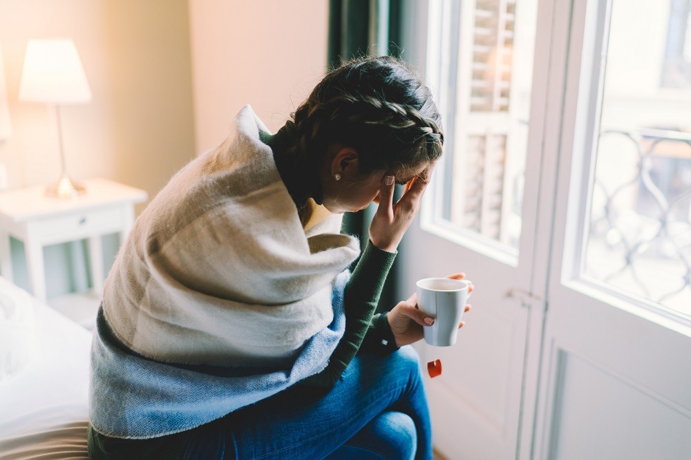 Woman sits on her bed and holds her head as she suffers with a cold