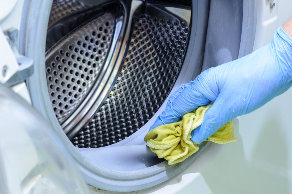 Someone in blue rubber gloves wipes inside of washing machine with a yellow cloth